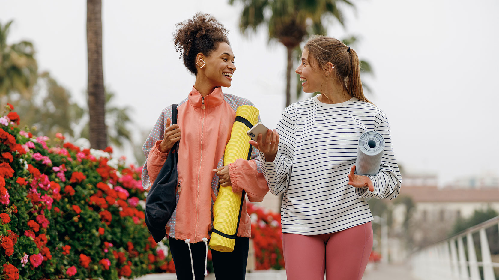 Two smiling female friend walking on morning city background with yoga mats after doing morning gym