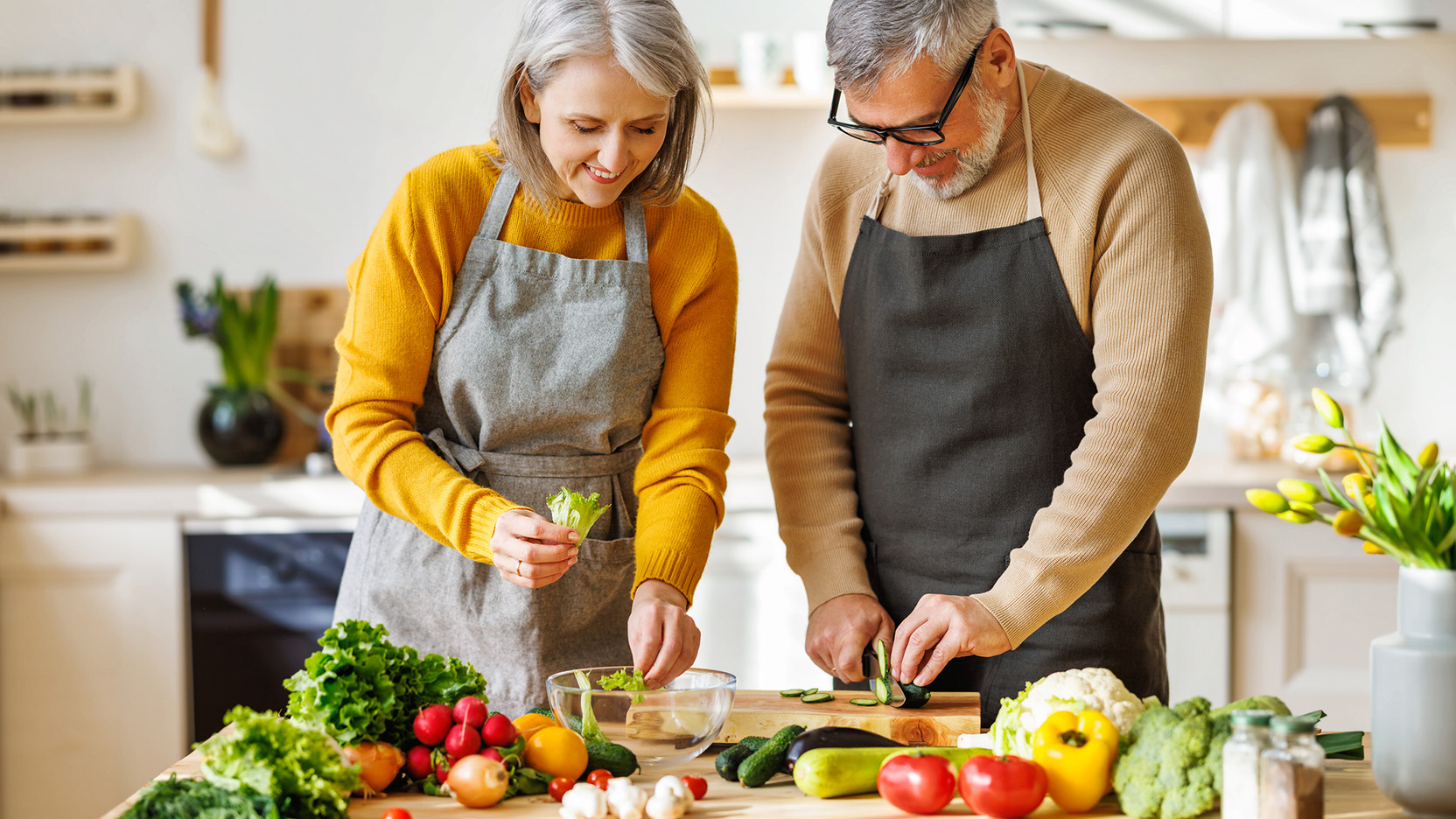 Happy elderly couple prepare vegetarian dinner together, chopping fresh colorful vegetables.