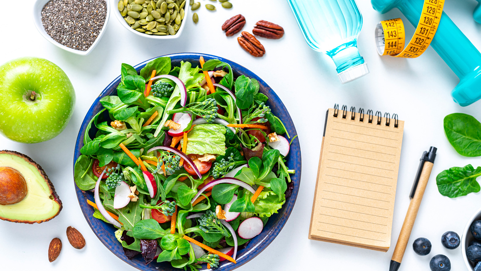 Overhead view of a healthy salad plate shot on white background. Water bottle, dumbbells and a tape measure are at the top right. A note pad with copy space is at the right. Multi colored fresh fruits, vegetables, seeds and nuts are around the salad plate.
