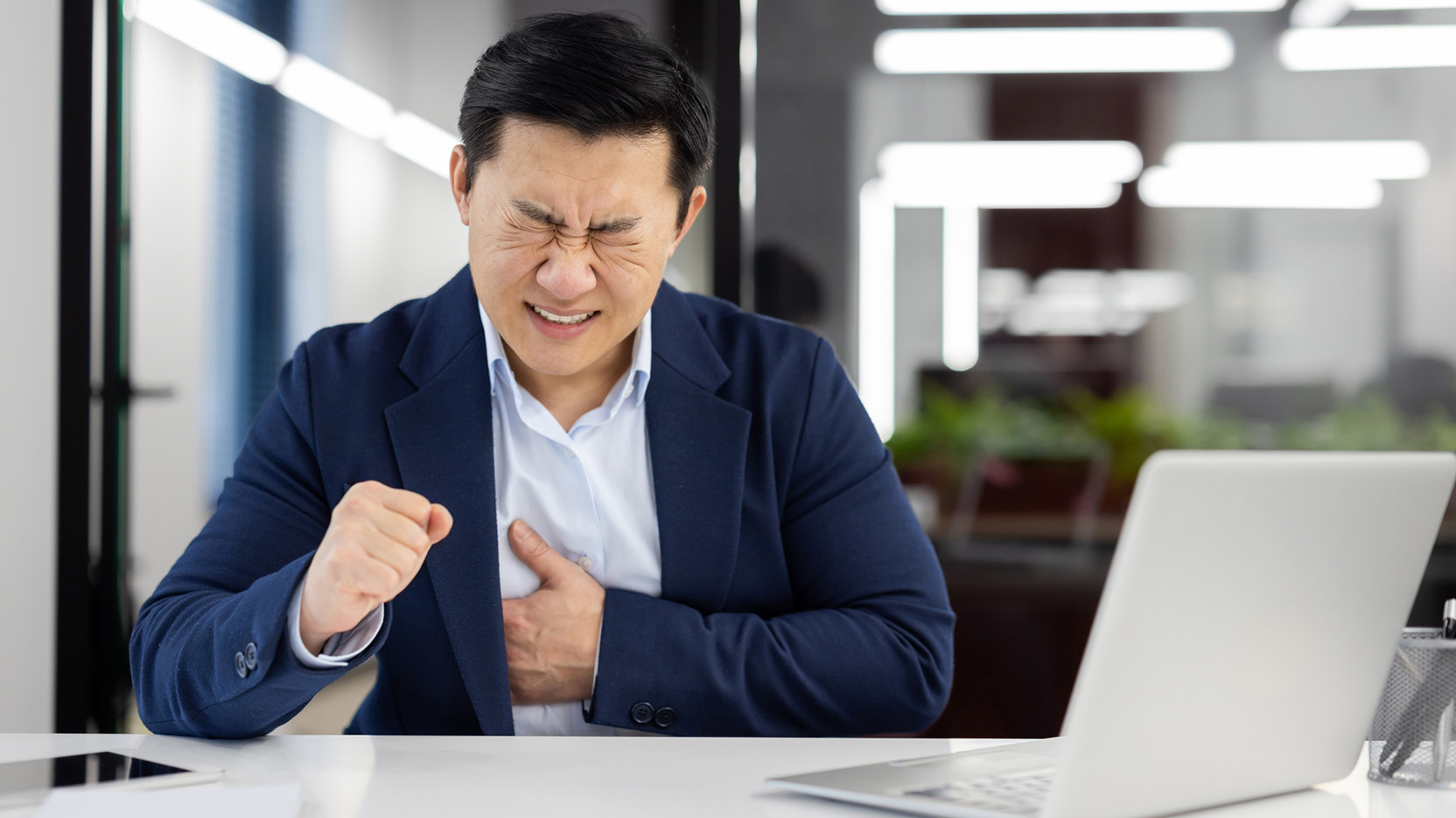 Heart pain in businessman, chest pain in asian man at workplace inside office, mature boss in business suit holding hands on chest, sitting at table with laptop.