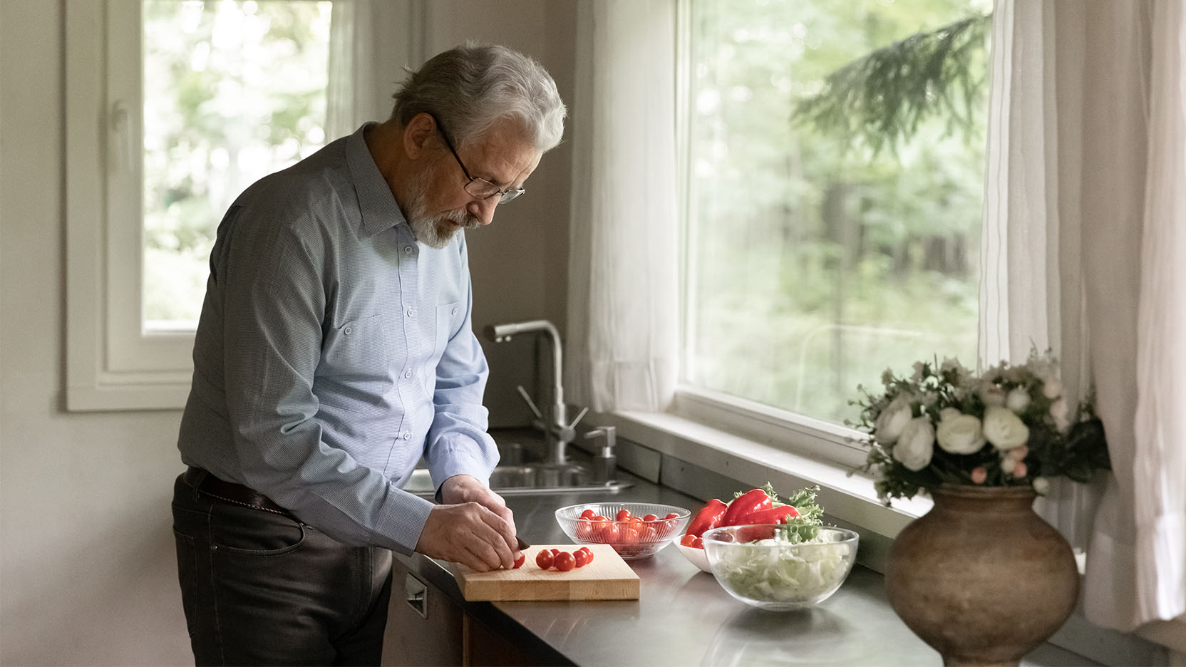 Older man cuts vegetables for a salad by a kitchen window