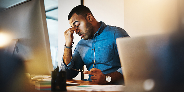 Professional Black man appears stressed while sitting in front of multiple computer monitors.