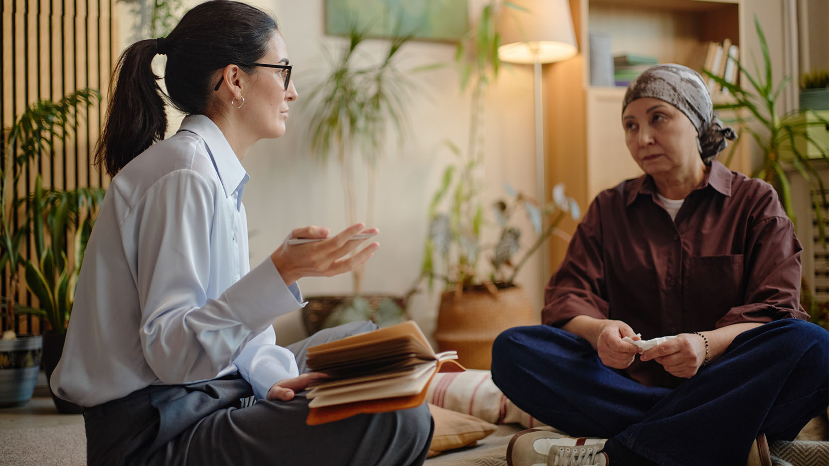 Two women, one wearing a headscarf, engage in conversation while seated on a living room floor.