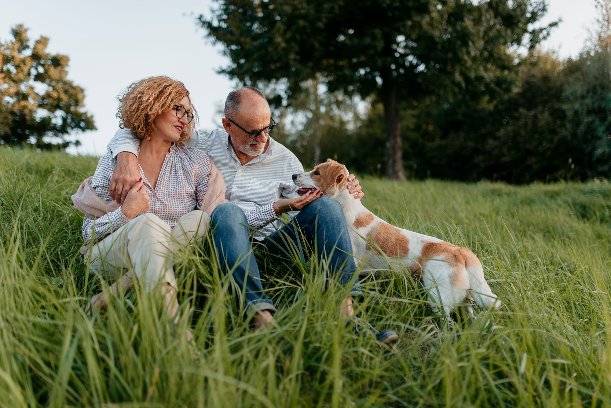Affectionate older couple sits outdoors in the grass with their dog 
