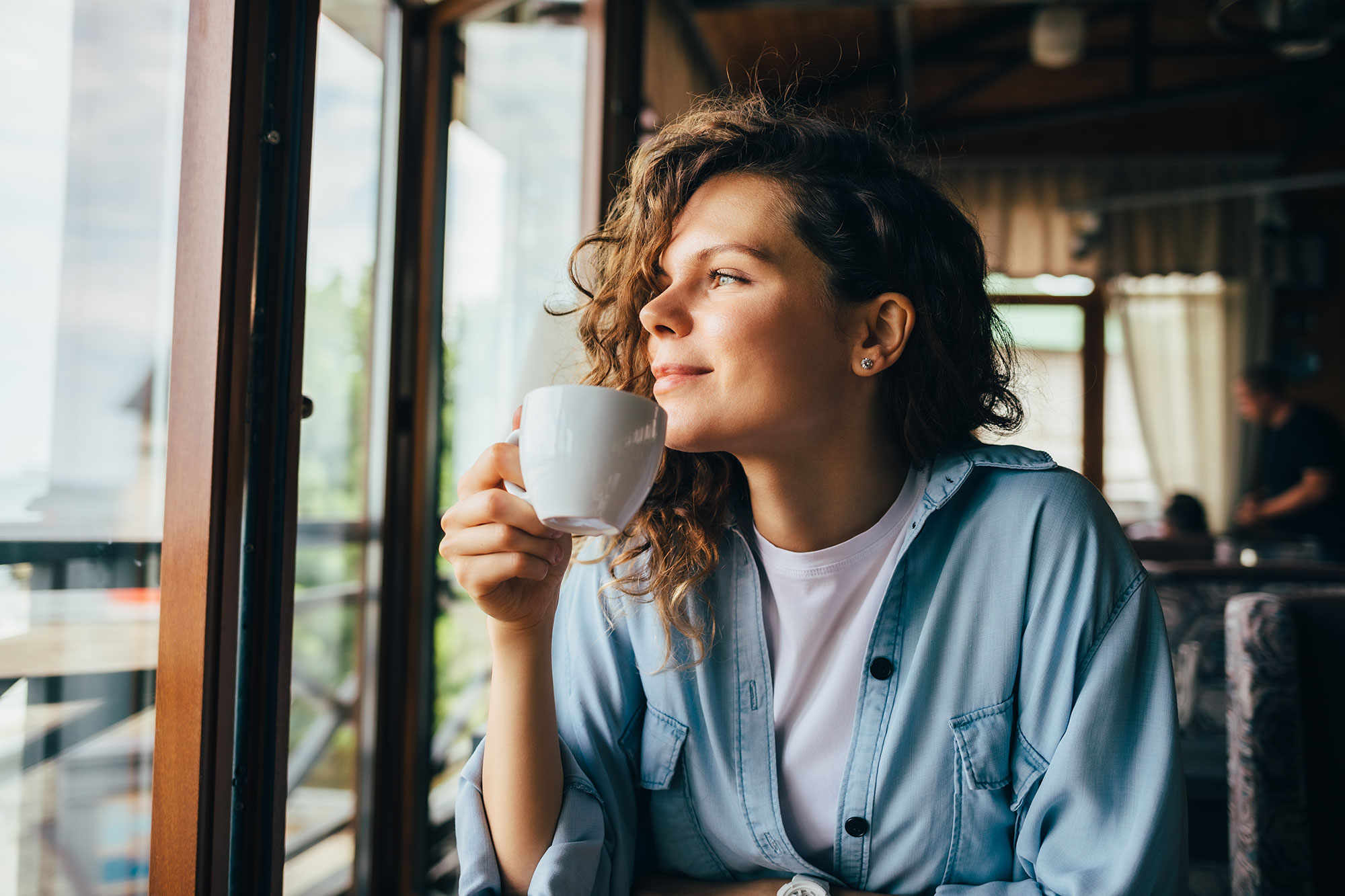 Smiling young woman looks out a window while drinking coffee