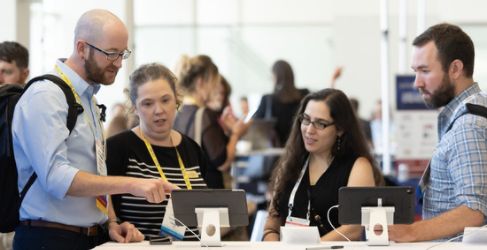 Group of four people stand around a laptop on a table and discuss what is on the screen.