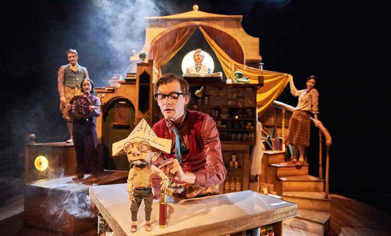 A man with eyeglasses manipulates a puppet who has round glasses and a paper hat. Four people stand on a set behind him, one holding a boat's wheel as though steering a ship.