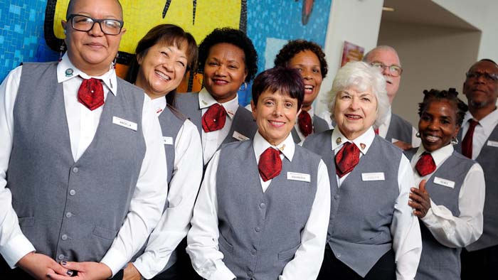 A group of about ten people, all wearing white shirts, gray vests, and red neckties, standing close together, facing the camera and smiling.