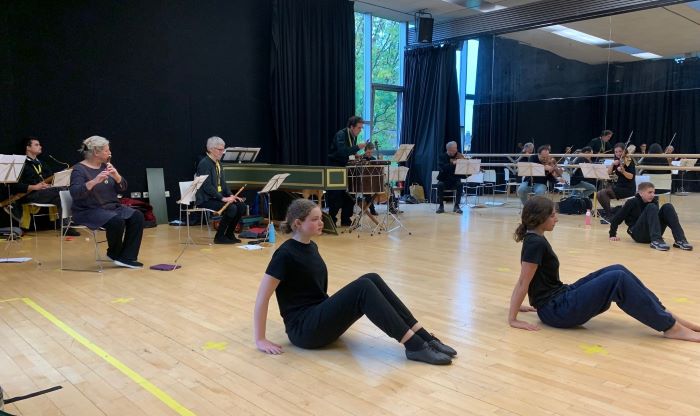 Students sitting on wooden floor rehearsing  with the Orchestra playing behind.