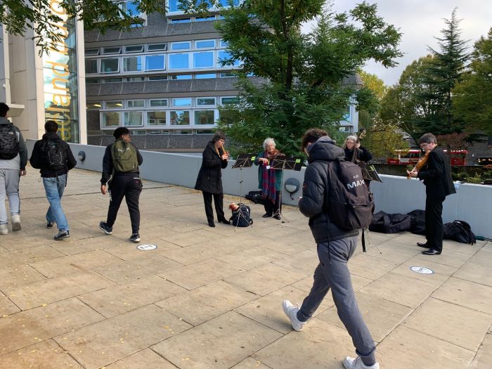 Students walking into school building with four members of an orchestra playing music to the right. 