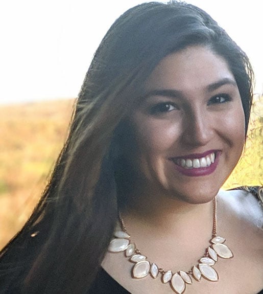 A person with long dark brown hair swept to one side, dark red lip color, and a a necklace seen from the shoulders up, smiling.