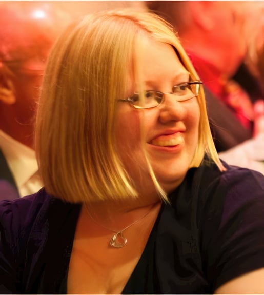 Caryl Jones smiling with medium length blonde hair and glasses, looking over the shoulder to the right