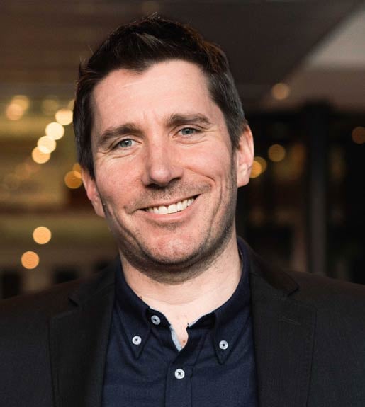 Headshot of a person with short brown hair and blue eyes, facing forward and smiling. Twinkling lights in the background.