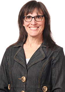A smiling woman with straight brown hair and glasses wearing a dark denim jacket