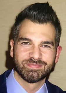 A man with brown hair and beard wearing a blue collared shirt and black jacket smiles directly into the camera