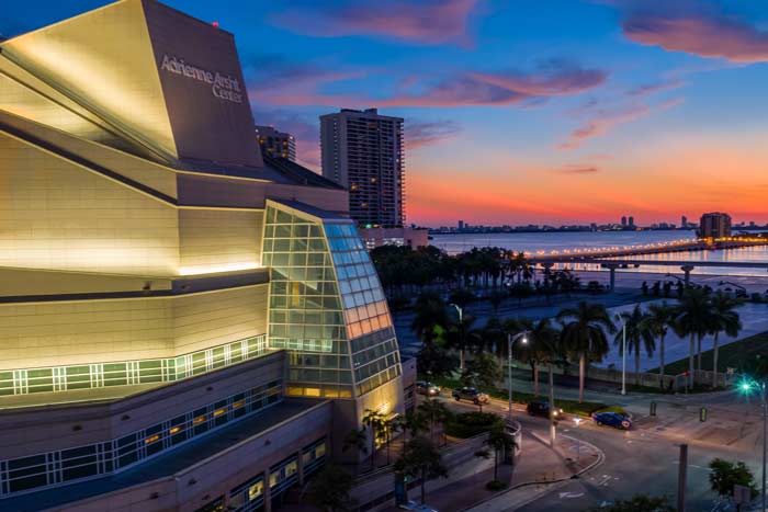 A large, white modern building, illuminated at early evening, with a body of water and a glowing sunset in the background.