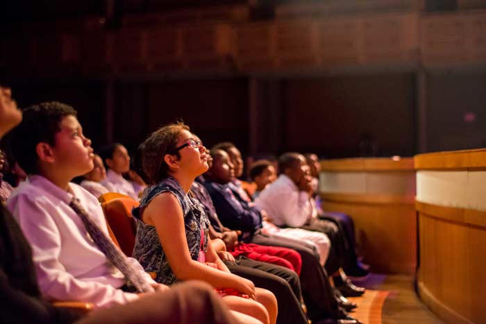 Young audience members sitting in a row of theater seats, seen from the side, looking up at a stage.