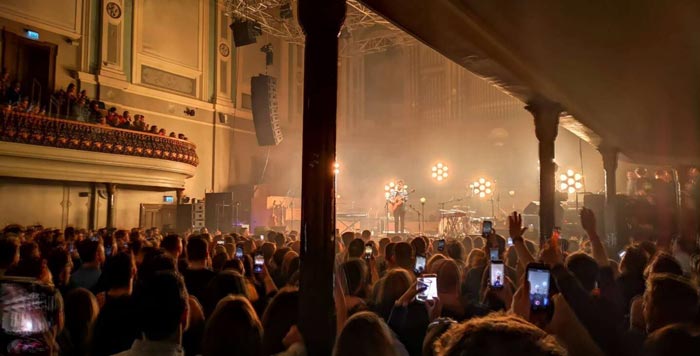 A performance hall filled with standing people, with fog in the air and lights on stage.