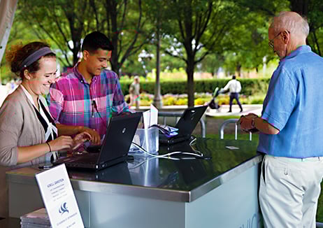 Two smiling people seen from the waist up, standing behind laptops on a counter. A person to the right is facing them as if to purchase or ask something.
