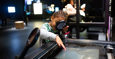 A young child looks through a magnifying glass at exhibit at We The Curious.
