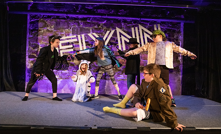 A group of children in costume performing onstage at the Flynn Center for the Performing Arts in front of a backdrop that reads 'Flynn.'