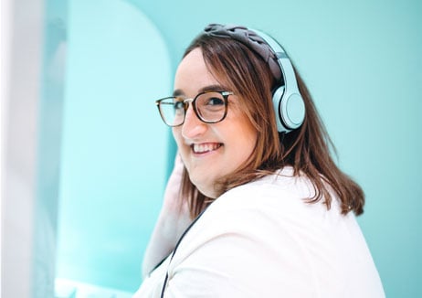 A woman wearing a headset, seen from the shoulders up, turning toward the camera and smiling. Aqua blue background.