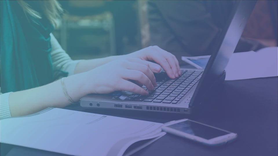 A photo showing a person's hands on the keyboard of a laptop. On the table next to the laptop are an open notebook and a mobile phone.
