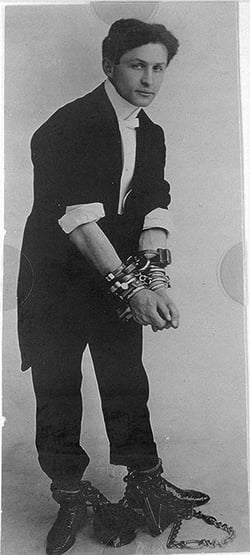 An old black-and-white photo showing a man with several pairs of handcuffs as well as leg shackles. 