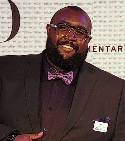 Quin Ansley, pictured smiling with dark glasses and a beard in front of a black and white sign, wearing a black suit and shirt with a bright purple bowtie.