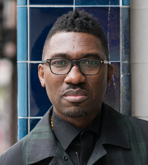 Kwame Kwei-Armah pictured from the shoulders up standing outside on a sidewalk while staring straight into the camera, wearing a black jacket and glasses