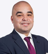 Photo of a man looking forwarding and smiling. He is wearing a dark blazer and purple tie and is seen from the shoulders up.