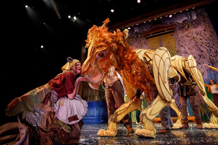 An actress leans her face close to a large, intricate puppet of a lion operated by three people