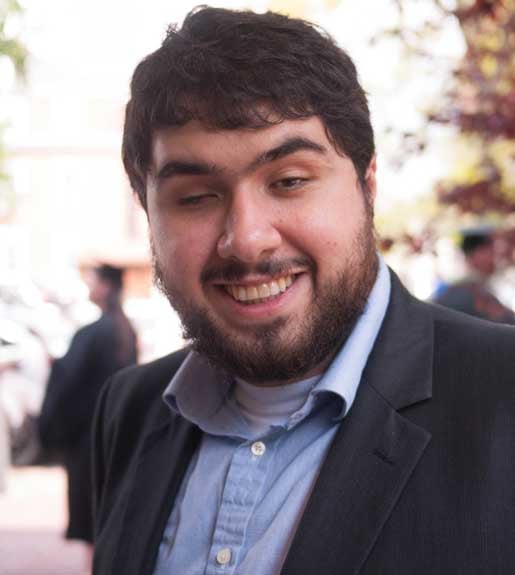 A man with dark brown hair, a beard, and a mustache. He is wearing a blue button-up shirt and a dark jacket, and is standing outdoors.