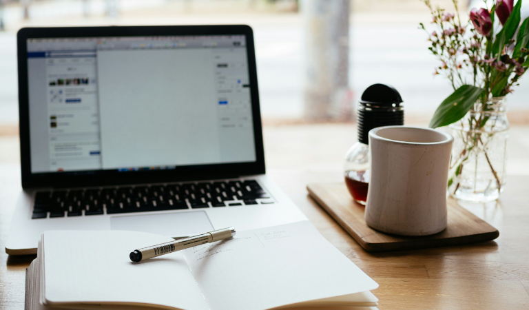 Photograph of open laptop on a desk with pen, notebook and mug
