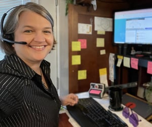 Person smiles at the camera from home office desk, wearing a head set.