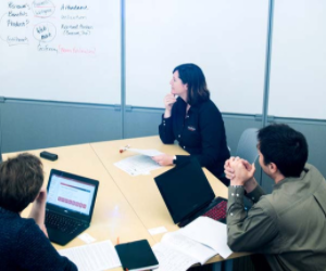 Three Tessitura team members sit around table with laptops looking up at whiteboard during meeting.