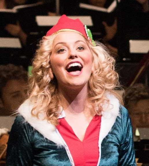 A woman with curly blond hair wearing an elf costume and singing. She is seen from the torso up.