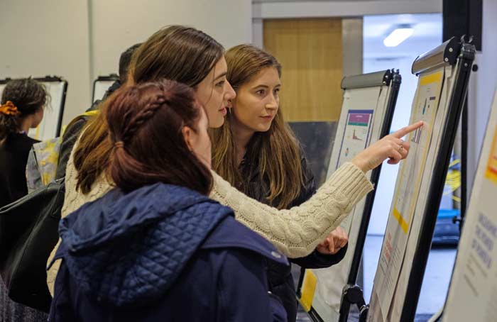 Three people standing and looking at a display on an easel. One person is pointing at something on the paper.