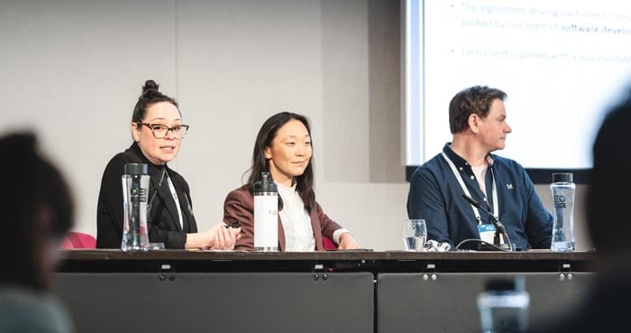 Three panelists sit side-by-side at a long table. The corner of a large screen is behind them.
