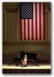Yo-Yo Ma sits on a chair in the middle of a stage holding a cello. Behind him is a black piano and an American flag hangs on the wall