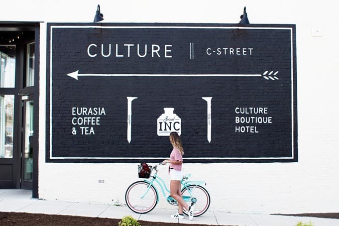 A woman rides a bicycle past a wall painted with a large arrow and the words Culture Boutique Hotel