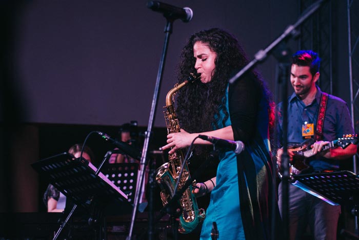A woman plays a saxophone on a stage. Behind her, a man plays an electric guitar
