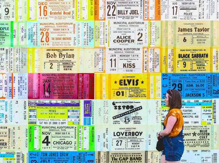 A woman walks past a wall covered in a collage of larger-than-life concert tickets for shows including Elvis Presley, Bob Dylan, Alice Cooper, and James Taylor