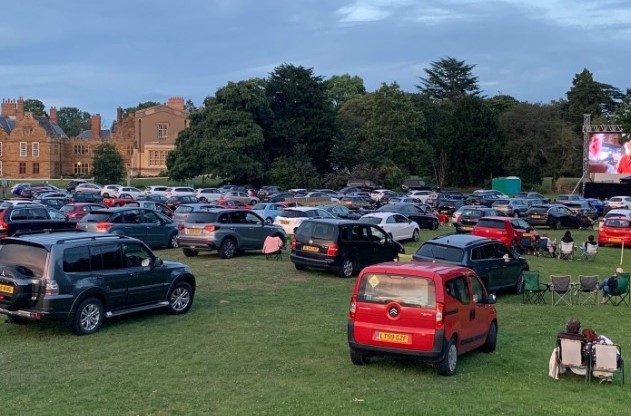 Cars parked on grass in front of the Delapré Abbey, with a an outdoor screen to the right of the frame.