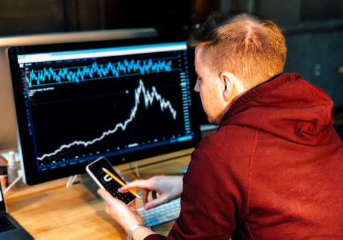 Person in red sweatshirt looking at phone with image of stock market on computer in the background. 