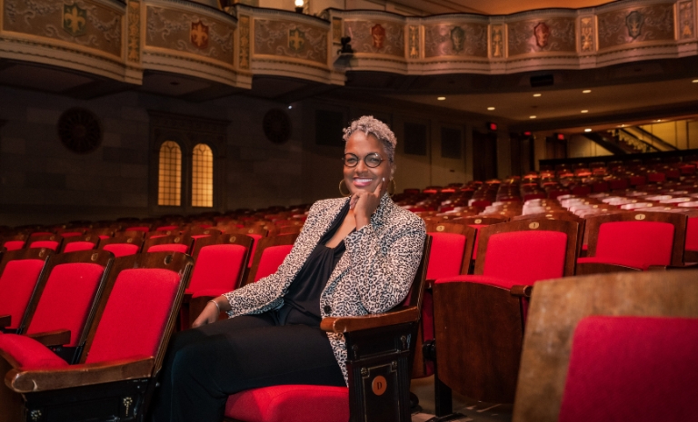 Sharon sits in the middle of a theater auditorium of red seats, smiling at the camera with chin resting on hand.