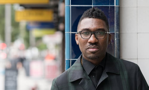 Kwame Kwei-Armah pictured from the shoulders up standing outside on a sidewalk while staring straight into the camera, wearing a black jacket and glasses