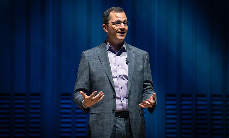 Andrew Recinos, a man with dark hair and glasses wearing a blue suit, presents in front of a dark blue background on a conference stage.