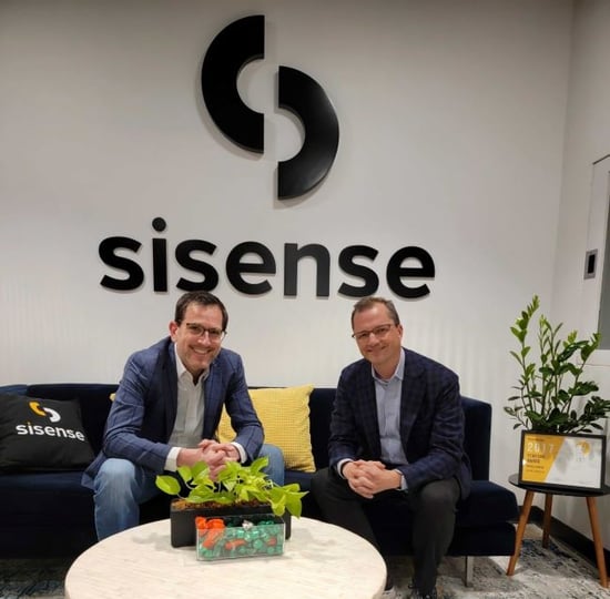 Amir and Andrew sitting on a couch with Sisense logo on the wall behind them and white coffee table in the front.