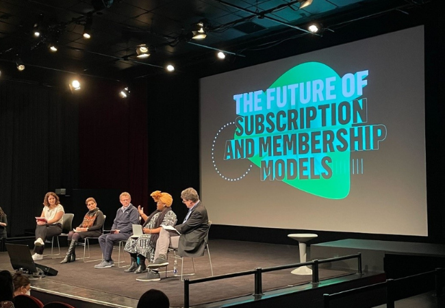 Speakers seated on stage in front of a screen with the text ' The Future of Subscriptions and Membership Models'